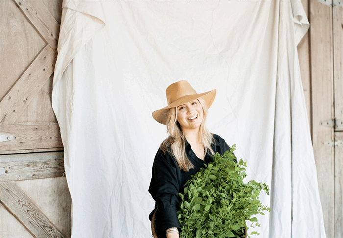 "Smiling woman in a wide-brimmed hat holding a basket of fresh green herbs, seated against a rustic wooden barn door and fabric backdrop."
