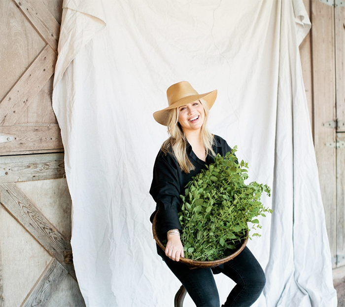 "Smiling woman in a wide-brimmed hat holding a basket of fresh green herbs, seated against a rustic wooden barn door and fabric backdrop."