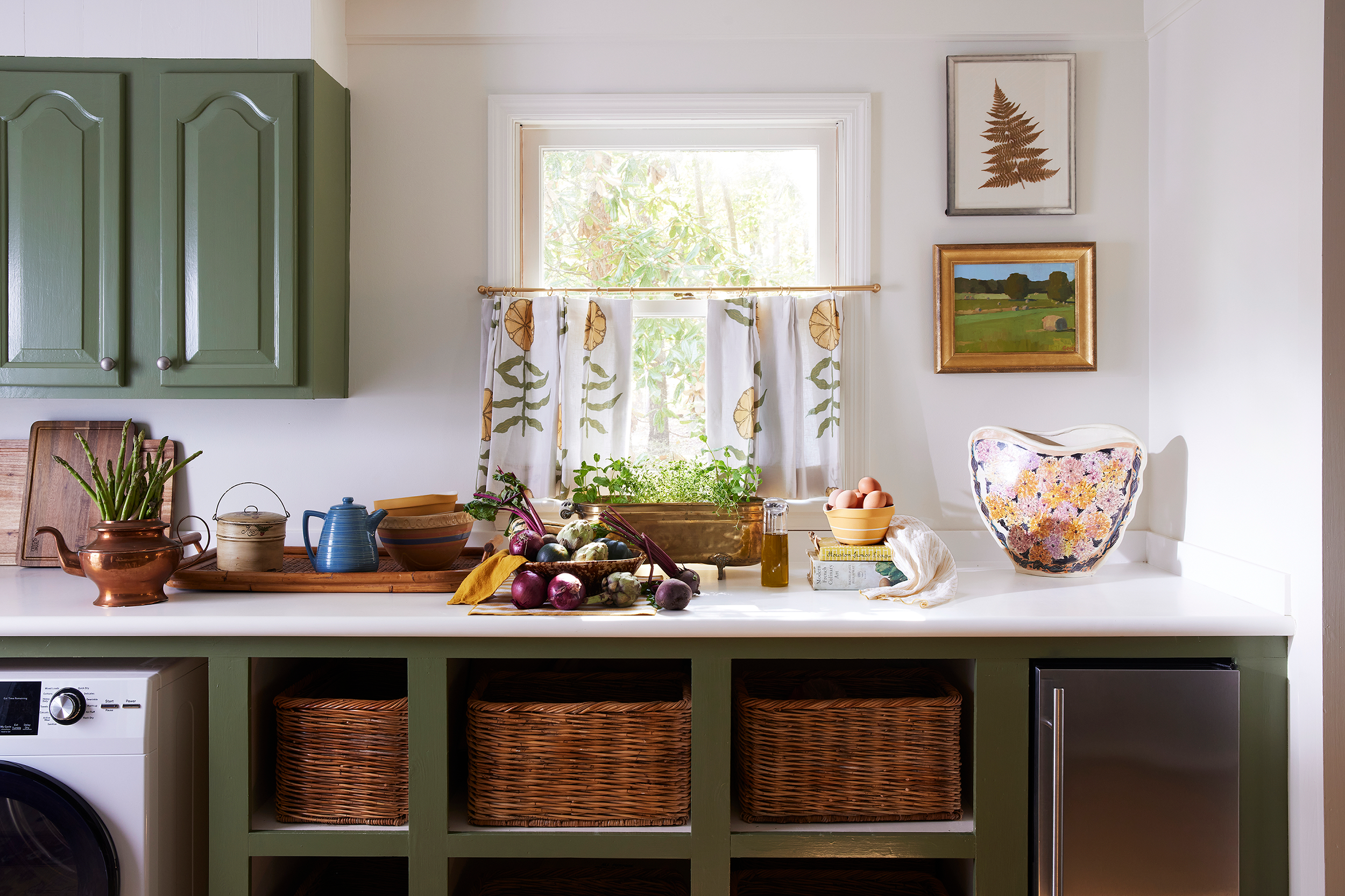 Green cabinetry beneath a window with printed café curtains, open wicker storage baskets, and a countertop. Styled with garden vegetables and a floral pitcher on display at a Lake Martin home. 