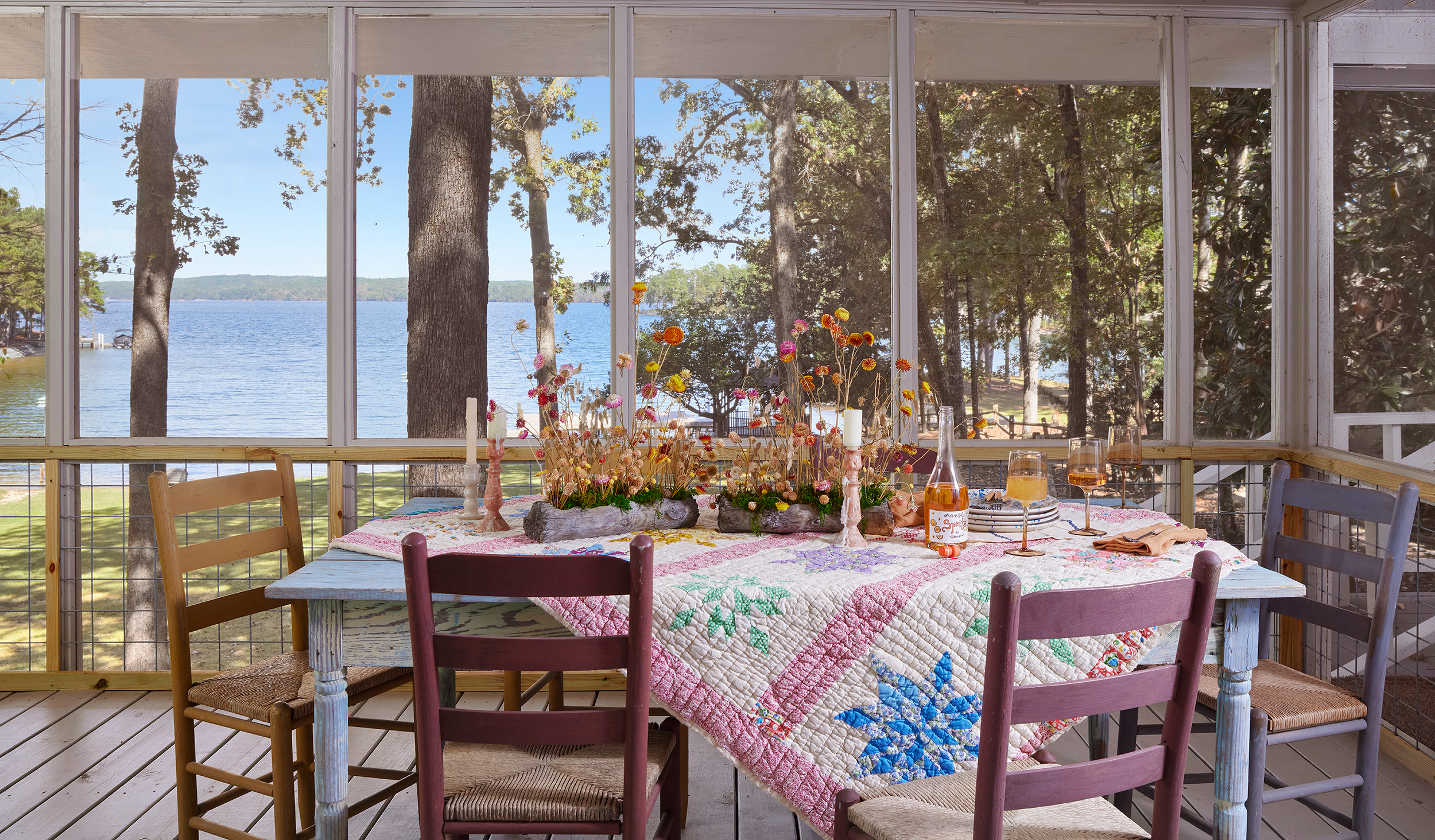 Screened porch dining table set with dried floral arrangements, candlesticks, and a vintage quilt tablecloth, overlooking Lake Martin through the trees.