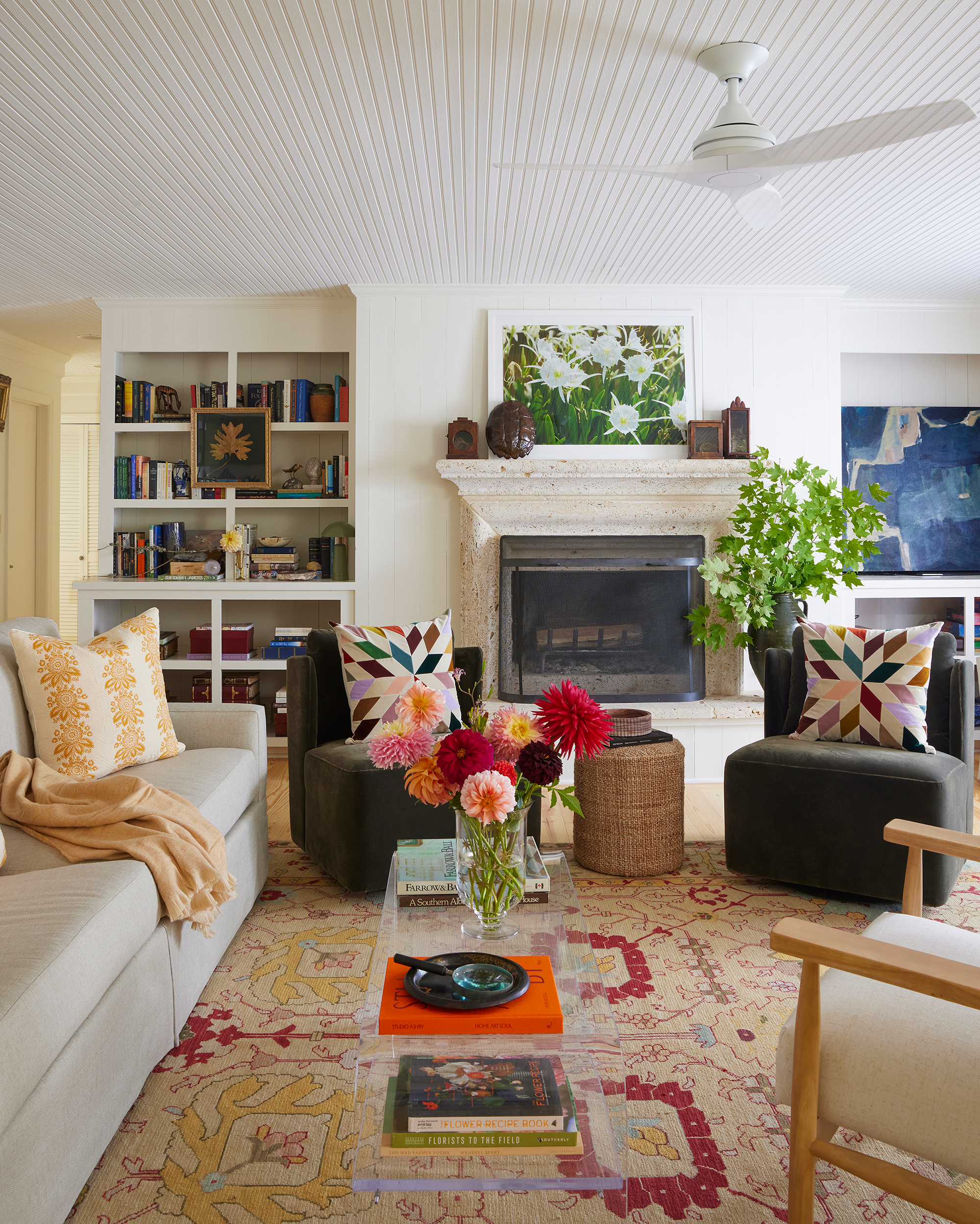 iving room with cream sofa, two olive-green swivel chairs, colorful quilted throw pillows, a stone fireplace, and a vase of fresh dahlias on a glass coffee table at a Lake Martin lake house. 