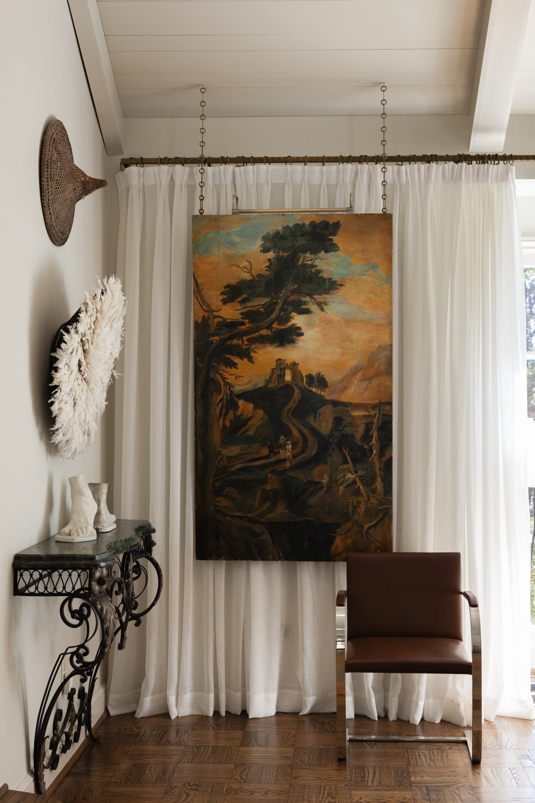 Large antique landscape painting hung from brass chains before sheer drapery in the living room of a Mountain Brook home
