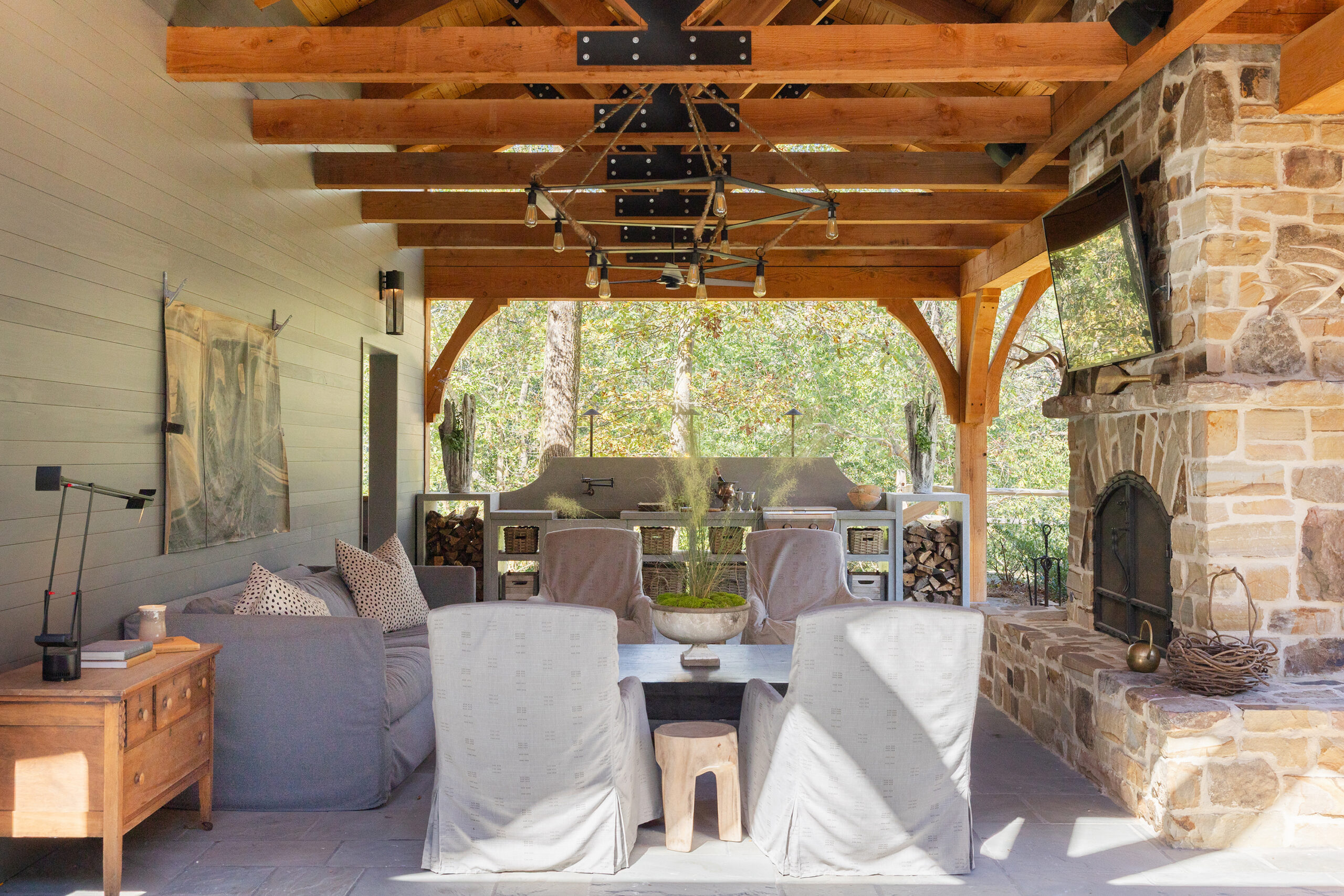 Covered outdoor living room in a renovated Mountain Brook 1950s ranch home featuring a double-sided stone fireplace, timber trestle ceiling, and custom concrete kitchen.