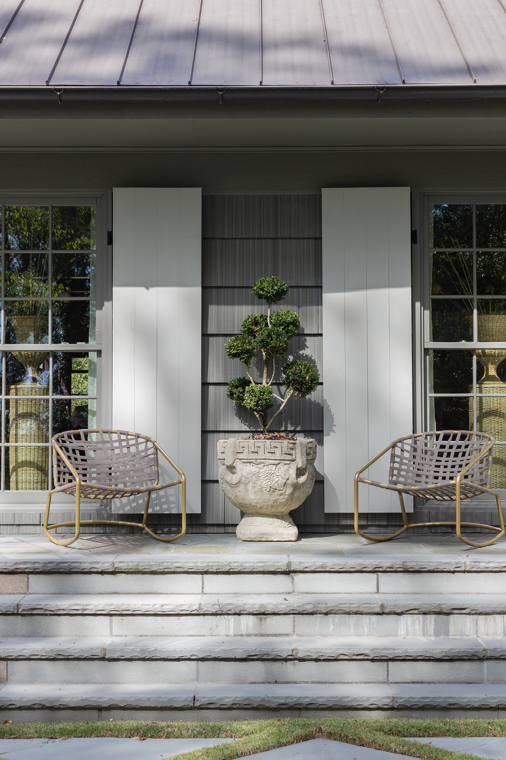 Front porch of a Mountain Brook cedar shake ranch featuring two gold-framed rocking chairs flanking a vintage cast-stone urn with topiary.